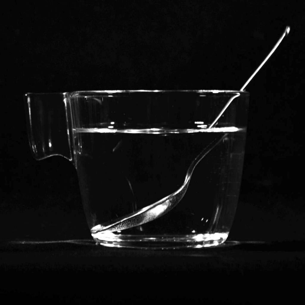 Ein Wasserglas, in dem sich ein Löffel befindet, vor schwarzem Hintergrund. An der Wasseroberfläche sieht es so aus als würde der Löffel einen Knick haben.