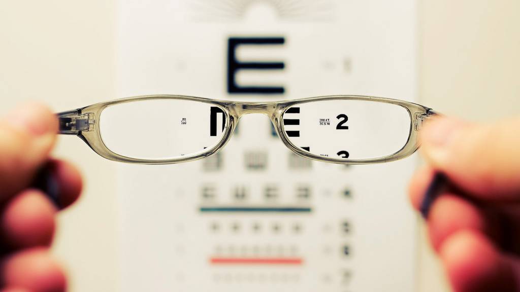 Zwei Hände halten eine Brille ins Bild. Im Hintergrund hängt ein Sehtest an der Wand. Die Zahlen und Buchstaben auf dem Sehtest sind nur unscharf, durch die Brillengläser jedoch scharf zu erkennen.