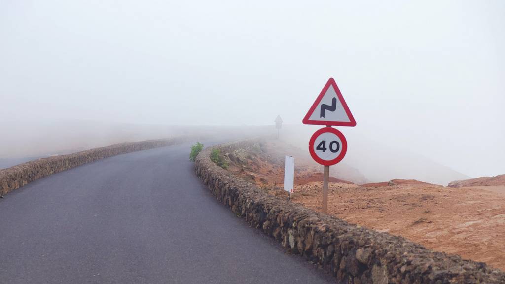 Eine Straße im Nebel. Im Vordergrund Verkehrsschilder, die auf eine scharfe Kurve und das Tempolimit 40 hinweisen.