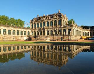 Blick auf den Westflügel des Dresdner Zwingers. Ein prunkvolles Gebäude im Barockstil. Im Vordergrund ein künstlicher Teich.