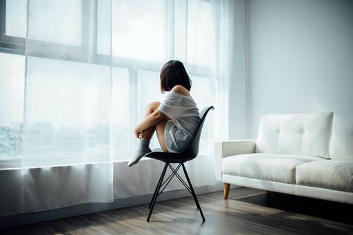 Eine Frau sitzt auf einem Stuhl in einem großen Raum und schaut aus dem Fenster. Andere Menschen sind nicht zu sehen.