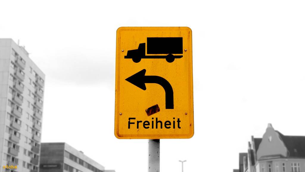 Ein Straßenschild zeigt an, dass LKW nach Links abzubiegen haben. Darunter hat jemand das Wort 